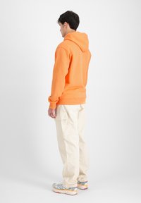 Sudadera naranja con bolsillo canguro, corte relajado, combinada con pantalones cargo beige. Los zapatos son de color azul claro con acentos texturizados y suelas multicolores.