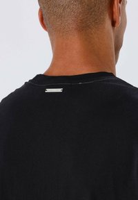 Camiseta de algodón negra con cuello redondo, que presenta una etiqueta plateada en la parte posterior del escote y una sutil textura en la tela.
