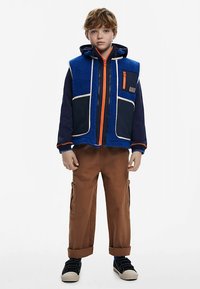 Modrá fleecová vesta s tmavomodrými rukávmi, oranžovým zipsom a detailmi. Hnedé kapsáčové nohavice s bočnými vreckami a zavinutými lemy. Čierne tenisky.