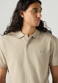 Beige polo shirt i mjukt tyg, med klassisk krage, två knappar och en diskret broderad logotyp på bröstet.