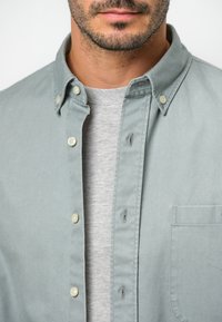 Camicia button-down verde chiaro con una tasca sul petto, colletto abbottonato e texture morbida, indossata sopra una t-shirt grigia a girocollo.