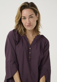 Lila Bluse mit Rundhalsausschnitt, drei dekorativen Knöpfen und 3/4-Ärmeln im Glockenstil. Bietet Falten-Details und eine weiche Stofftextur.