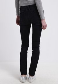 Jeans skinny de mezclilla negra con una cintura de altura media, que cuentan con bolsillos traseros y una textura suave; combinados con zapatillas metálicas plateadas.