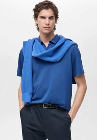 Sininen polo-paita, jossa on tekstuuri-neuleita harteilla, yhdistetty tummiin housuihin. Sujuva kangas, rento muotoilu ja puhtaat linjat.