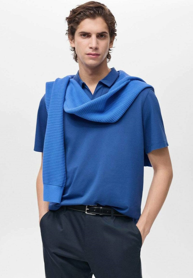 Sininen polo-paita, jossa on tekstuuri-neuleita harteilla, yhdistetty tummiin housuihin. Sujuva kangas, rento muotoilu ja puhtaat linjat.