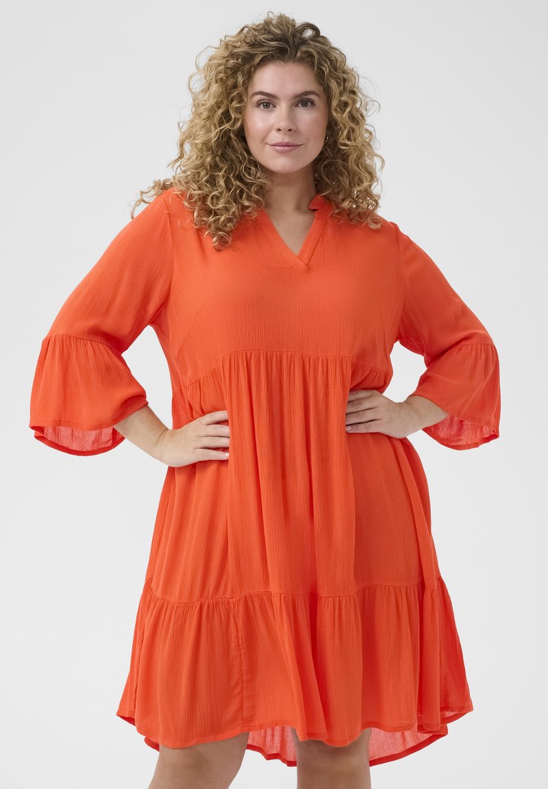 Kortsleeves, v-ringning, knädlång klänning i klar orange tyg med en texturerad yta och flerlagd fåll. Har lösa, utsvängda ärmar.