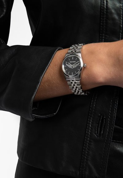 Srebrny zegarek ze stali nierdzewnej z okrągłą czarną tarczą, srebrnymi znacznikami godzin i bransoletą w formie ogniw. Noszony na nadgarstku obok czarnej skórzanej kurtki.