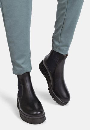Black leather ankle boots with chunky soles worn with light green pants, showing a heart detail on elastic side panels.
