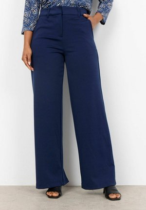 Femme portant un pantalon large bleu marine avec des passants de ceinture et des sandales à talons ouvertes noires, debout contre un mur blanc.