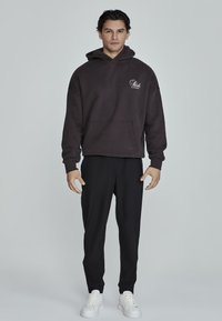 Hanorac gri închis cu buzunar frontal și logo alb, asortat cu pantaloni negri tapered și adidași albi, pe un fundal simplu.