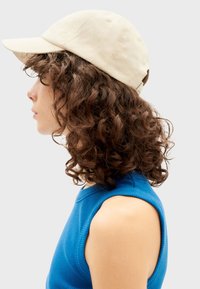 Beige katoenen pet met een gebogen vizier, zichtbare stiksels en een verstelbare strap, gecombineerd met een blauwe geribbelde mouwloze top.
