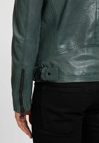 Groene leren jas met een gestructureerde afwerking, verticale stiksels, een zijrits en een verstelbare gesp in de taille. Zwarte broek zichtbaar.