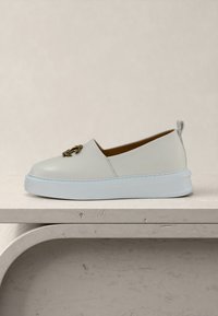 Κρεμ slip-on αθλητικό παπούτσι με λευκή παχιά σόλα και μεταλλικό διακοσμητικό έμβλημα στο πάνω μέρος, τοποθετημένο σε επιφάνεια με μπεζ υφή.