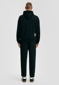 Zwarte hoodie met een trekkoordkap, ruime pasvorm en logodetail op de achterkant. Gecombineerd met tapse zwarte broek en casual schoenen.