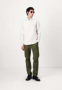 Camisa de botonadura en color beige claro con rayas sutiles, pantalones verdes y zapatos negros. Los accesorios incluyen gafas de sol. Apariencia limpia y moderna.