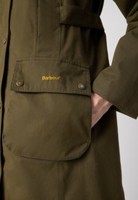 Olijfgroene katoenen jas met grote voorzakken, drukknoopsluiting en gouden "Barbour" logo-details. Zachte textuur, praktische ontwerp.