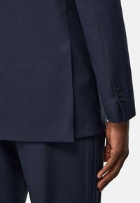 Costume bleu marine avec un motif texturé, des coutures visibles et des accents de poignets boutonnés. L'ouverture au dos et la coupe ajustée sont mises en avant.