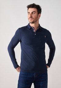 Marine langermet polo skjorte med knappestolpe, røde og hvite krageaccentene, og lomme på venstre bryst. Laget av glatt stoff.