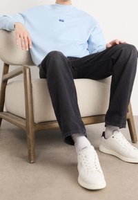 Felpa azzurra chiara con un piccolo logo, abbinata a jeans neri e sneaker bianche. L'individuo è seduto su una poltrona in tessuto chiaro.