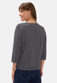 Kvinna som ler, iklädd en svart och vit randig topp med tre kvarts ärm och mörkblå jeans, stående mot en enkel bakgrund.