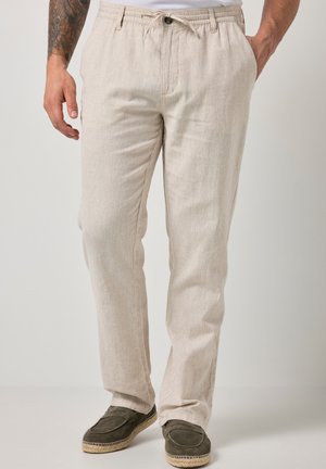 Mężczyzna w beżowych lnianych spodniach ze ściągaczem w pasie i ciemnych zamszowych butach wsuwanych, stojący na jednolitym tle.