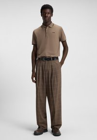 Hombre vestido con una camisa polo beige de Hugo, pantalones de cuadros marrones de pierna ancha, cinturón negro y zapatos marrones, de pie contra un fondo liso.