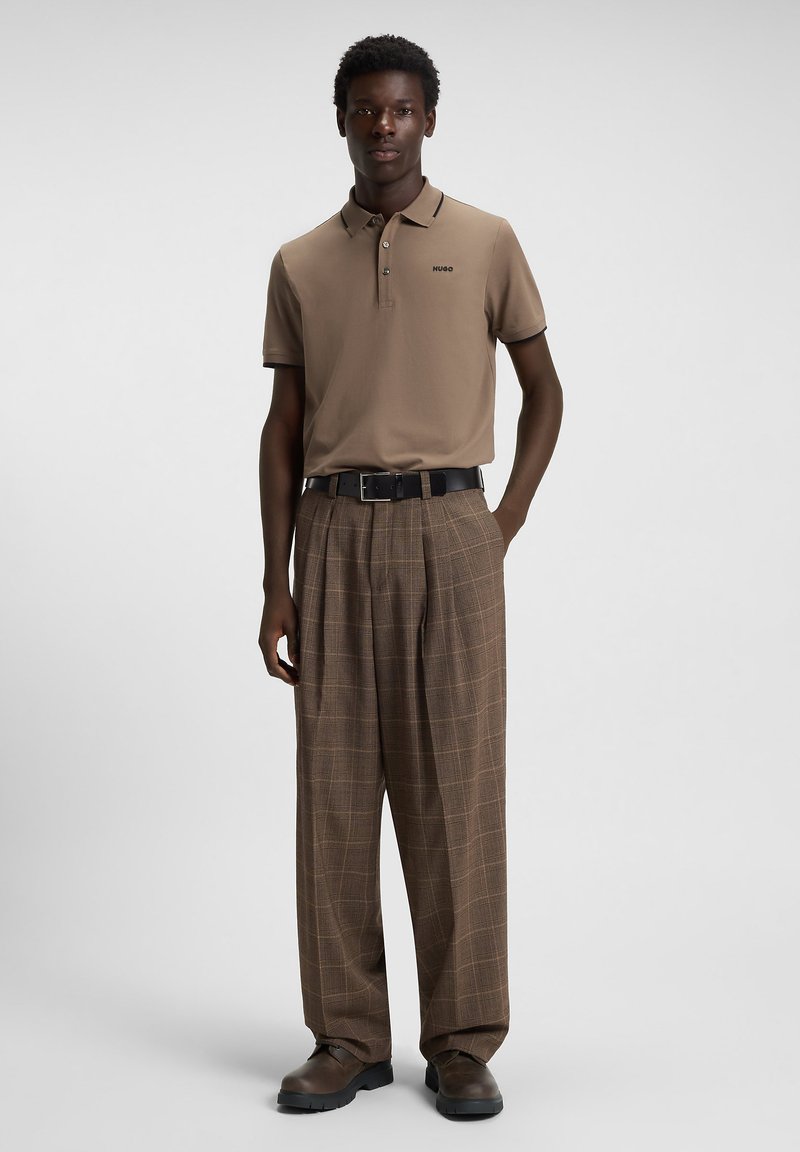 Hombre vestido con una camisa polo beige de Hugo, pantalones de cuadros marrones de pierna ancha, cinturón negro y zapatos marrones, de pie contra un fondo liso.