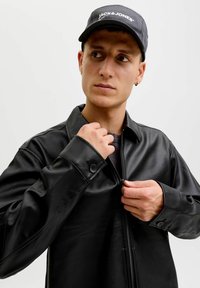 Schwarze Lederjacke mit durchgehendem Reißverschluss und Knopfmanschetten. Getragen über einem schwarzen Rundhals-Shirt, ergänzt durch eine Mütze.