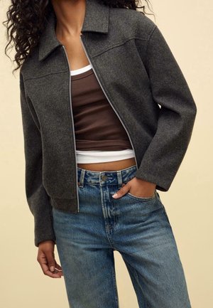 Kobieta ubrana w ciemnoszarą kurtkę zapinaną na zamek, na brązowym topie i niebieskich dżinsach, stojąca z jedną ręką w kieszeni na beżowym tle.