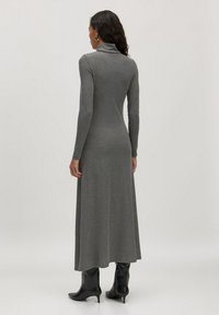 Femme aux cheveux bouclés portant une robe maxi à manches longues en turtleneck grise et des bottes à talons noires, se tenant de dos devant un fond uni.
