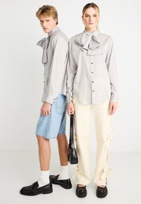 Ριγέ πουκάμισο με κουμπιά και μεγάλο φιόγκο σε γκρι και μπεζ χρώμα· συνδυασμένο με ανοιχτόχρωμο τζιν σορτς και κρεμ παντελόνια με διακοσμητικούς φιόγκους. Μαύρα παπούτσια.