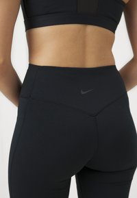 Leggings noirs taille haute avec une texture lisse, présentant un logo Nike noir subtil et des détails de couture à la ceinture. Vue arrière.
