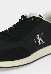 Μαύρο αθλητικό παπούτσι Calvin Klein με λευκό λογότυπο "cK" στο πλάι, μαύρα κορδόνια, λευκό επενδεδυμένο γιακά και υπόλευκη σόλα.