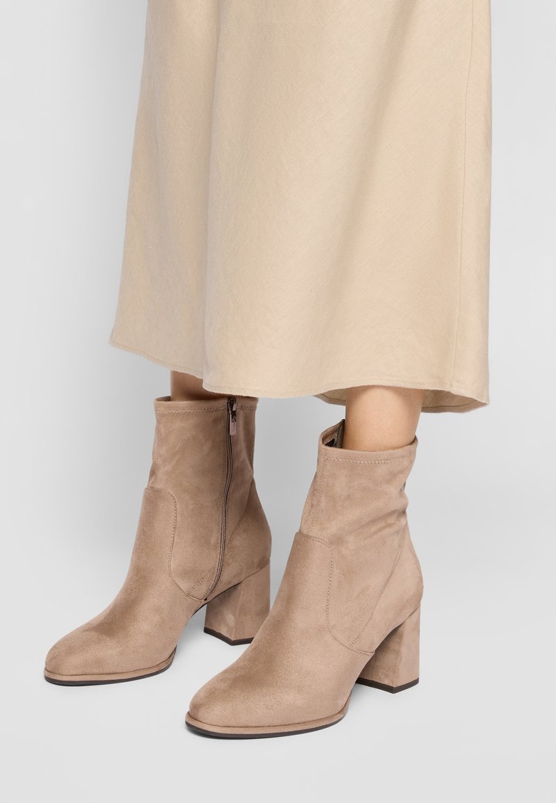 Bottes courtes en daim beige avec talons carrés et fermeture éclair latérale, portées avec une jupe midi beige clair, debout sur un sol gris clair.