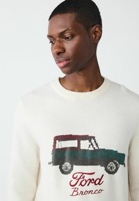 Kreem värvi kootud kampsun, millel on tumedates toonides Ford Bronco graafika koos punaste ja roheliste aktsentidega. Ümmargune kaelus, tekstureeritud kangas.