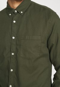 Homme portant une chemise boutonnée vert olive avec une poche poitrine et des boutons blancs, visible du cou à la taille.