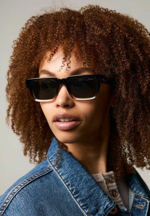 Zwarte en doorzichtige oversized zonnebril met een dik frame, gedragen door iemand met krullend, bruin haar, gekleed in een spijkerjack.