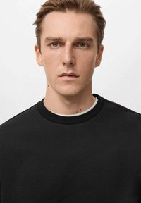 Mladý muž s krátkymi svetlohnedými vlasmi má na sebe čierny tričko s okrúhlym výstrihom cez biele tričko, pozerá priamo do kamery, s neutrálnym výrazom.