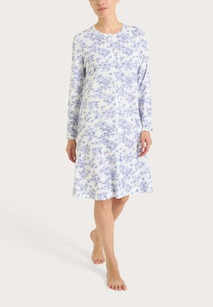 Donna con indosso una camicia da notte bianca fino al ginocchio con motivi floreali e farfalle blu, in piedi a piedi nudi su uno sfondo chiaro.