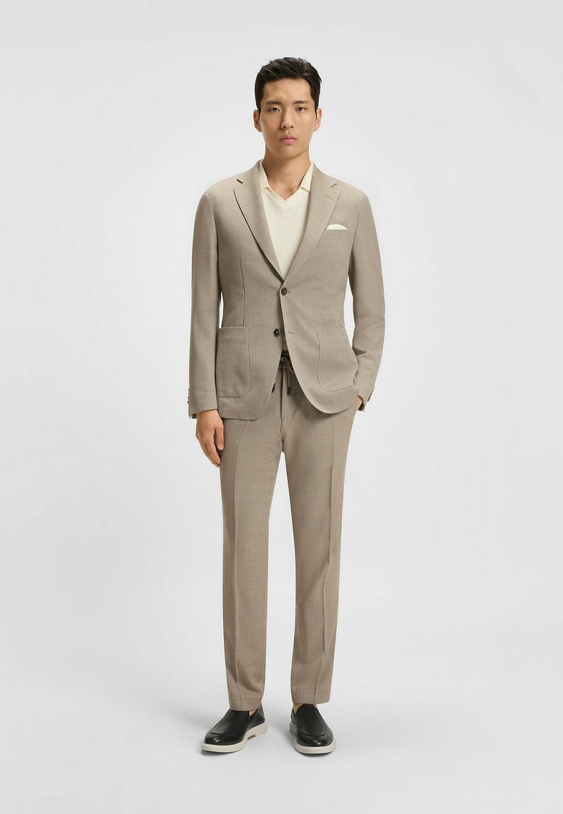 Costume beige avec un tissu texturé, veste à un bouton, revers crantés, deux poches avant, associé à un haut en tricot léger et des chaussures noires.