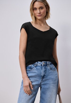 Nainen, joka pukeutuu mustaan, hihattomaan toppiin ja vaaleansinisiin farkkuihin, ja pitää beigeä laukkua kädessään yksiväristä vaaleaa taustaa vasten.