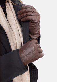 Brown leather gloves with a textured pattern and stitched details, worn over a beige scarf and black coat. Hands grasping a coat pocket.
