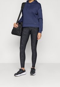 Felpa con cappuccio blu navy con una tasca frontale, abbinata a leggings neri con un delicato motivo a foglie. Scarpe da ginnastica nere e borsa a tracolla nera.