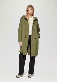 Oliwkowy parka z kapturem ze sznurkiem, przednimi kieszeniami i zasuwanym zamkiem, nałożona na kremowy sweter i czarne spodnie. Srebrne sneakersy.