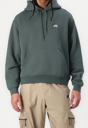 Osoba ubrana w ciemnozieloną bluzę z kapturem Nike SB z przednią kieszenią oraz beżowe spodnie cargo Nike z bocznymi kieszeniami na zamek.