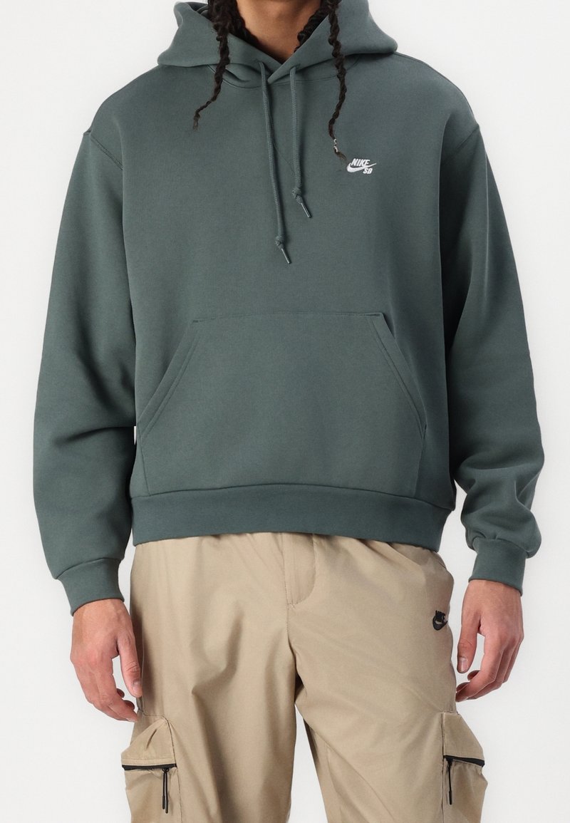 Personne portant un sweat à capuche Nike SB vert foncé avec une poche avant et un pantalon cargo beige Nike avec des poches à fermeture éclair sur les côtés.