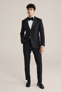 Mladenič oblečen v črno tuxedo s belo srajco, črno metuljčko in črnimi oblečnimi čevlji, stoji pred enobarvnim ozadjem.