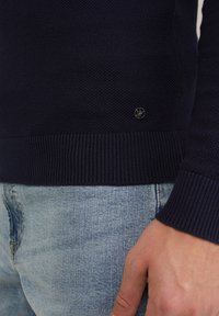 Dunkelblauer Strickpullover mit gerippten Bündchen und Saum, mit einem kleinen Logo-Detail nahe dem unteren Rand. Über hellblauen Jeans getragen.