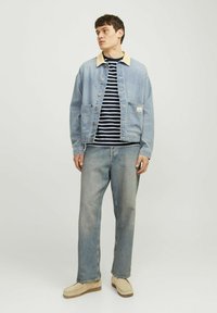 Chaqueta de mezclilla con cuello beige, cierres de botones, dos bolsillos; camisa a rayas azul marino y blanca debajo; pantalones de mezclilla claros; zapatos beige.