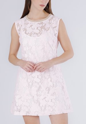 Femme portant une robe sans manches en dentelle florale rose clair avec un col rond et une bordure beige, debout les mains légèrement jointes.